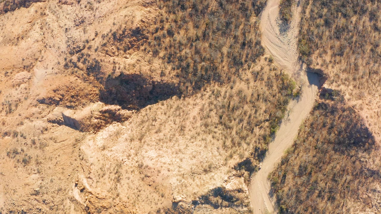 Overhead frame from Cabo Pulmo, Baja California Sur showing a pale dirt road curving between tan rocky hills and dry brush, with no visible people.