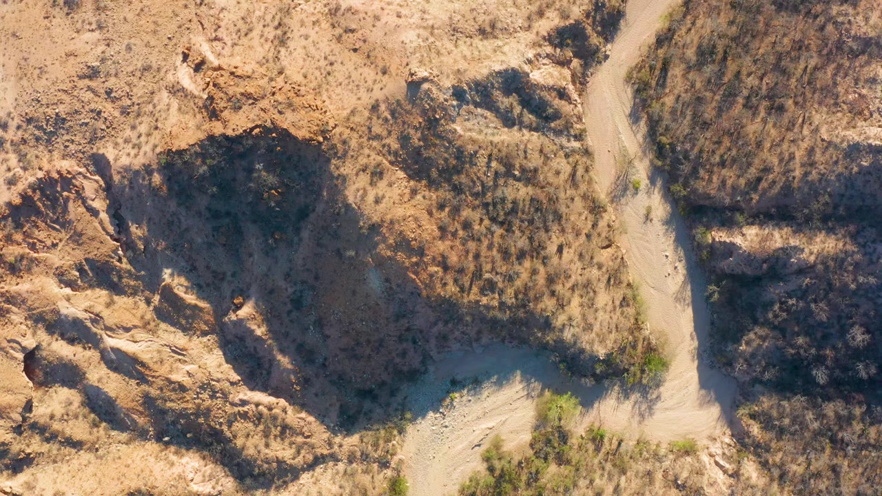 Aerial view in Cabo Pulmo, Baja California Sur, with the dirt road bending through dry brush and shadowed rocky cuts, with no visible people.