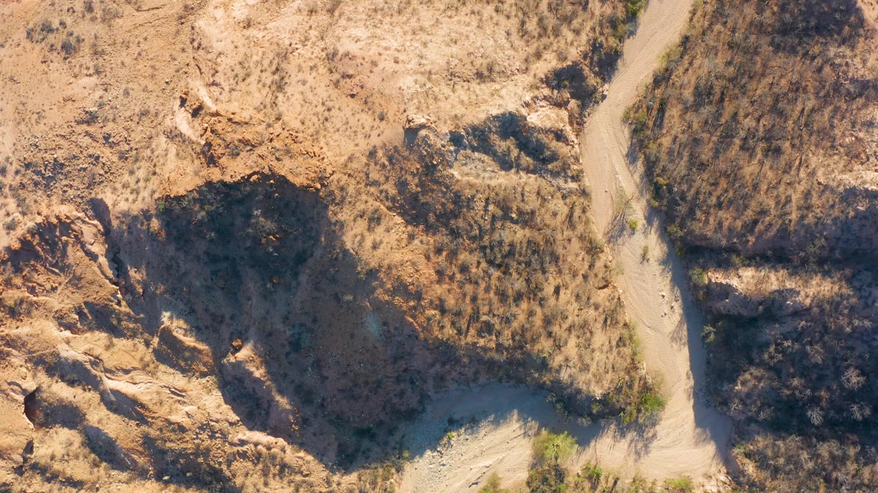 Overhead frame from Cabo Pulmo, Baja California Sur, where a narrow dirt road cuts through beige rocky slopes and dry scrub, with no visible people.