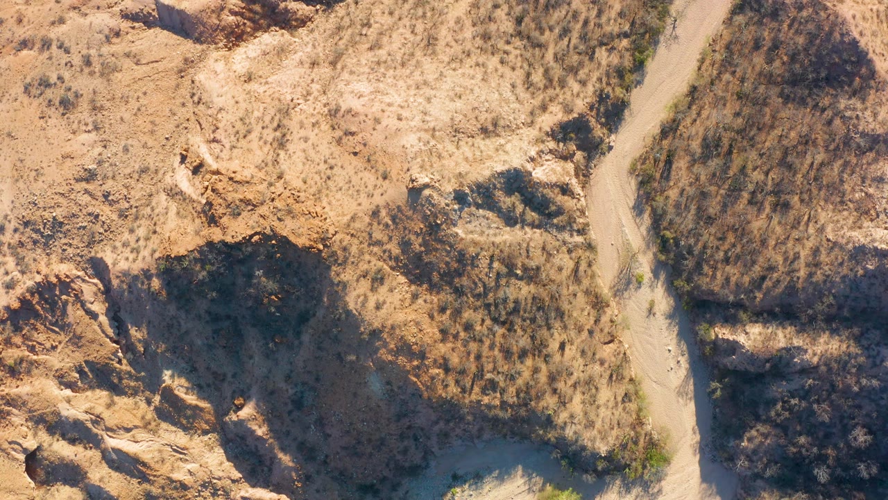Cabo Pulmo, Baja California Sur aerial frame showing the road alongside a dry wash, eroded rock, tan grasses, and deep shadow, with no visible people.