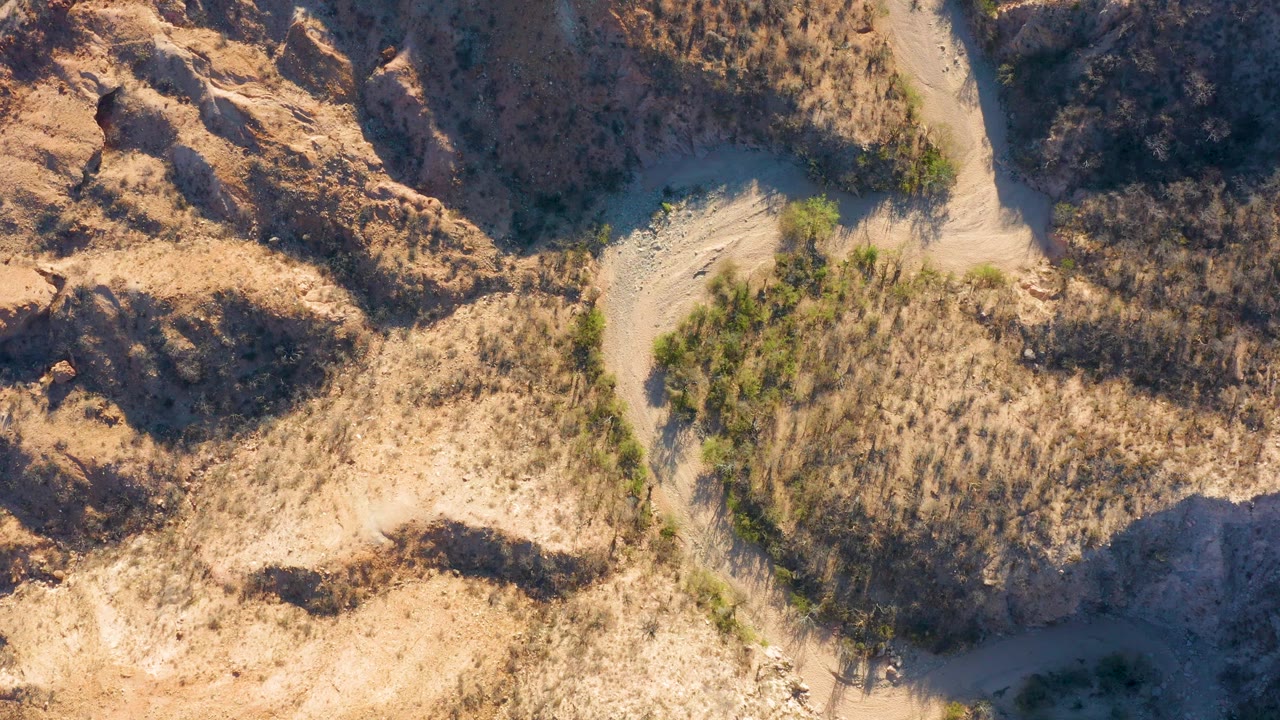 Aerial frame over Cabo Pulmo terrain with the dry wash running down the center, scrub-covered slopes on both sides, warm tan soil, and no visible people.