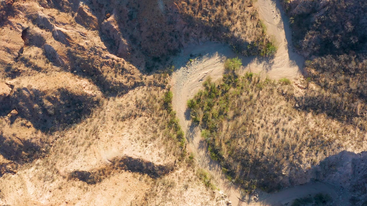 Aerial frame over Cabo Pulmo showing a branching dry channel, rough tan rock forms, sparse green vegetation, dry grass texture, and no visible people.