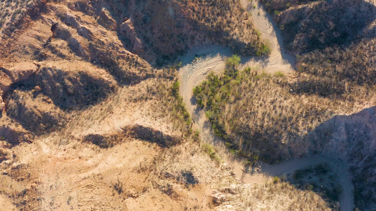 Top-down frame from Cabo Pulmo, Baja California Sur showing the pale road crossing near dry brush, rugged brown ridges, and a darker shadowed slope, with no visible people.