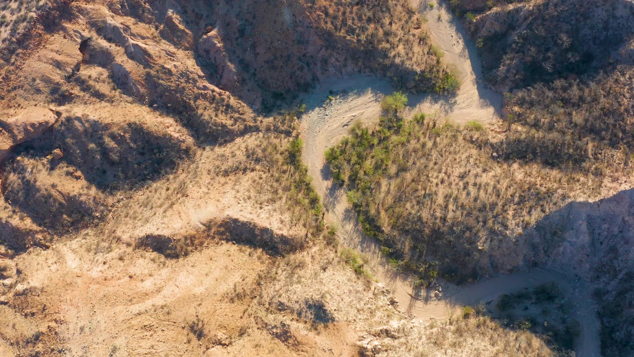 Overhead frame from Cabo Pulmo, Baja California Sur where the dirt road curves along a brushy rise, surrounded by tan earth, rocky banks, and no visible people.