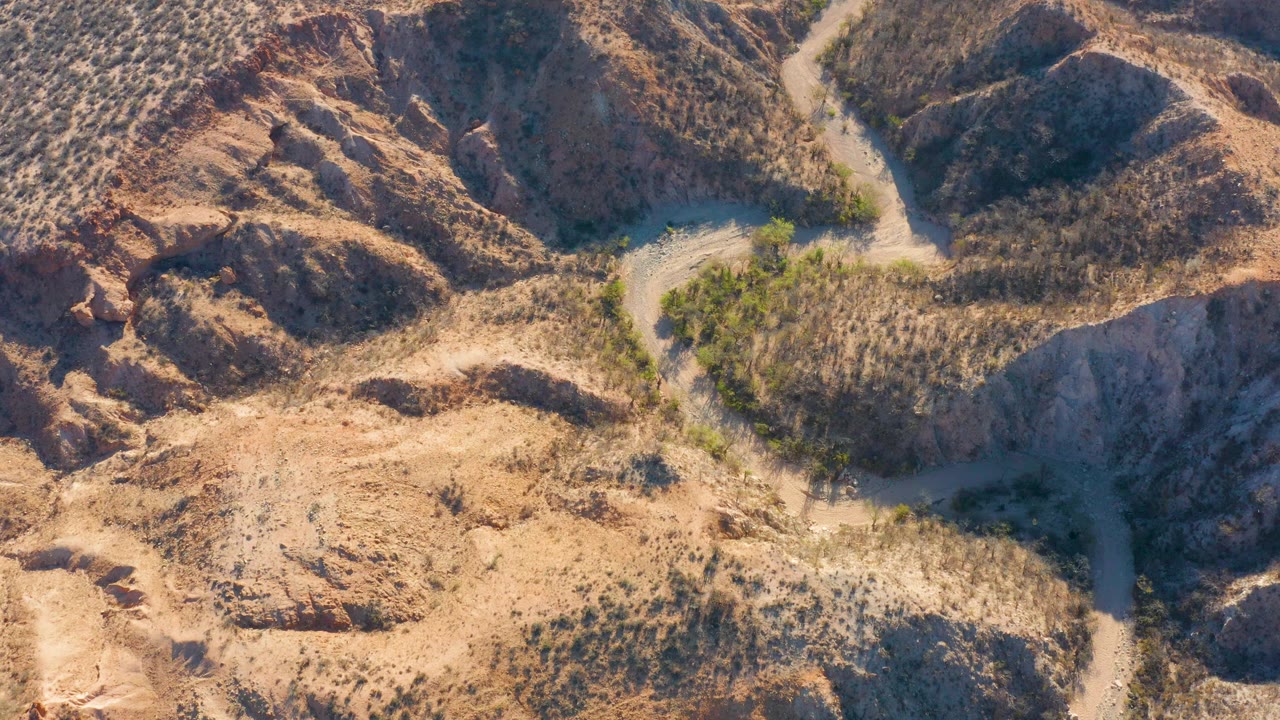 Opening aerial frame over Cabo Pulmo, Baja California Sur, with a pale dirt road curving between tan scrub and reddish rocky hills, no visible people.