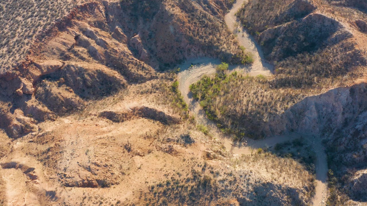Cabo Pulmo aerial view with a pale road crossing through dry grass, low shrubs, and rounded rocky hills under strong sunlight, no visible people.