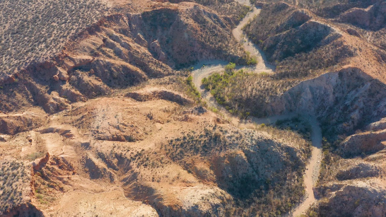 Cabo Pulmo desert terrain seen from above, with the dirt road shifting closer to center as it bends past dry brush and rocky slopes, with no visible people.