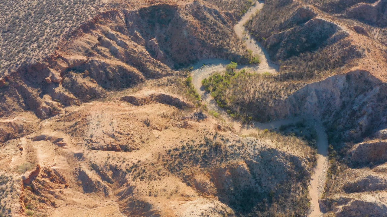 Aerial frame over Cabo Pulmo hills where the road bends between rounded tan slopes and darker scrub-covered gullies, with no visible people.