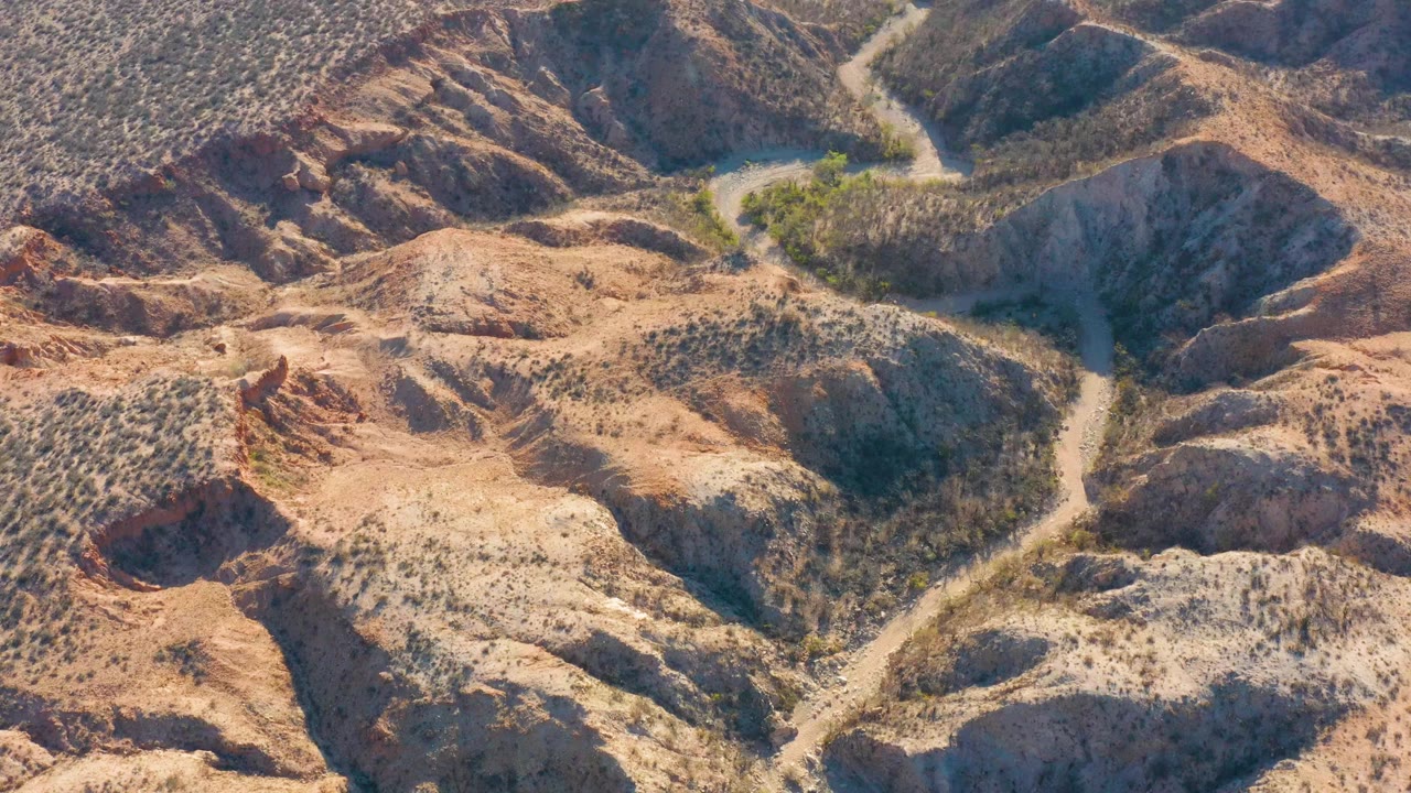 Aerial frame over Cabo Pulmo, Baja California Sur, showing a pale dirt road winding between tan rocky hills and sparse scrub, with no visible people.