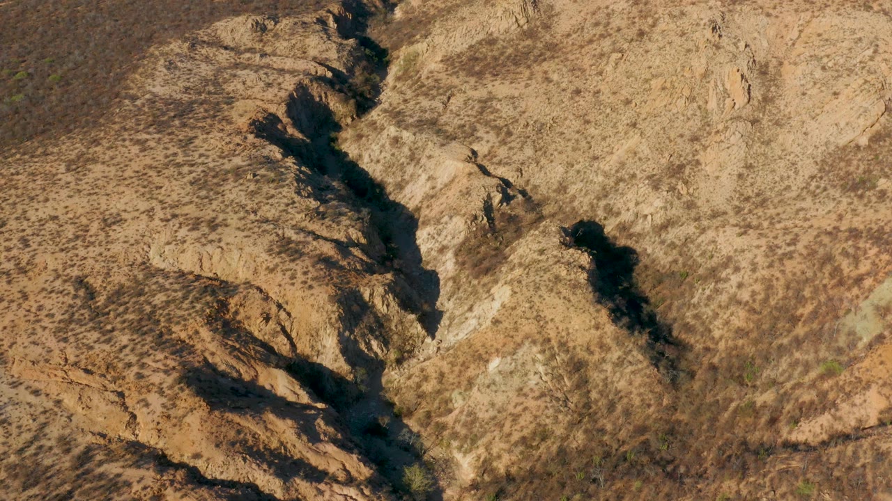 Overhead aerial frame from Cabo Pulmo, Baja California Sur showing a narrow dark water channel between beige rocky slopes and sparse scrub, with no visible people.