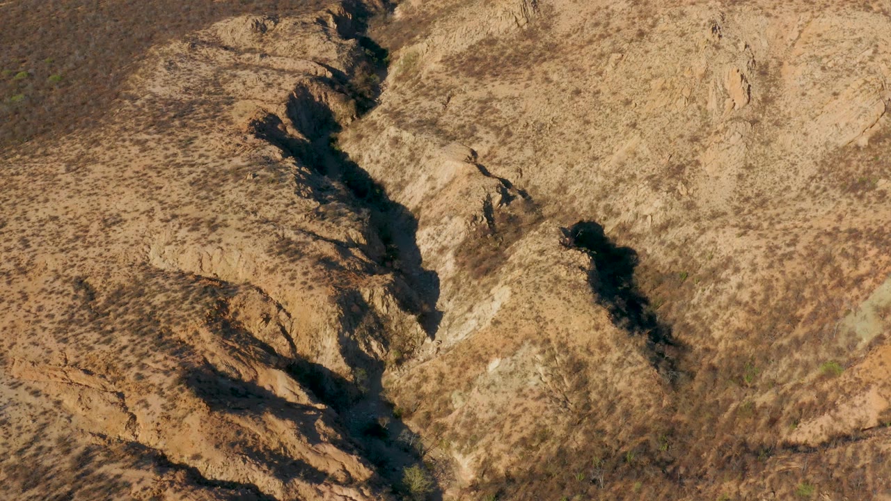 Aerial frame from Cabo Pulmo, Baja California Sur with the narrow channel slightly farther through the composition, bordered by tan ridges, dark shadow pockets, and no visible people.