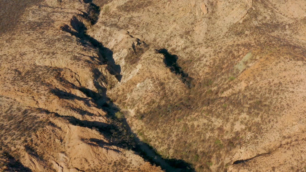 Aerial frame over Cabo Pulmo, Mexico showing a narrow dark water channel cutting diagonally through tan rocky terrain, with sunlit dry ground and no visible people.