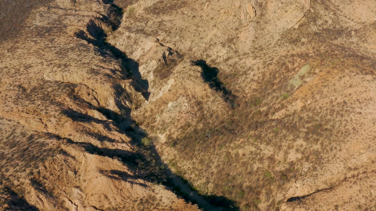 Overhead Cabo Pulmo frame with the water channel centered between shadowed rock cuts, dry tan slopes, scattered scrub texture, and no visible people.