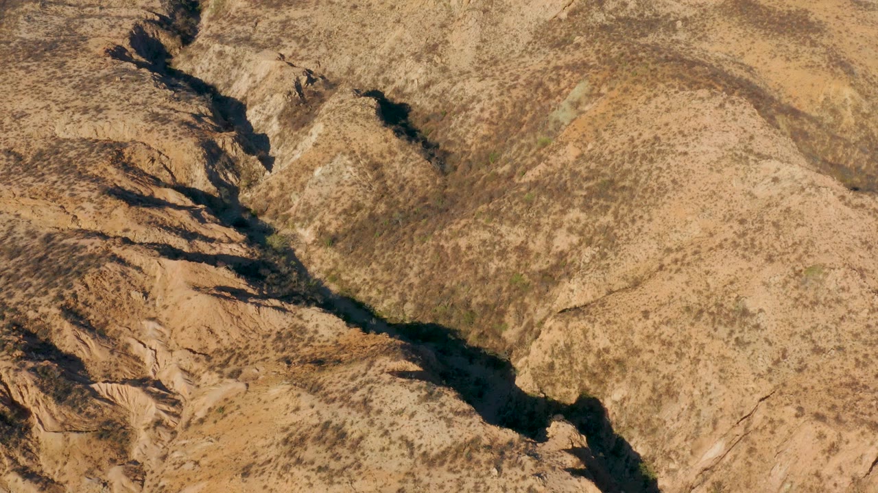 At 0:01, the aerial composition centers the winding wash between sunlit tan slopes and darker eroded rock bands in Cabo Pulmo, with no visible people.