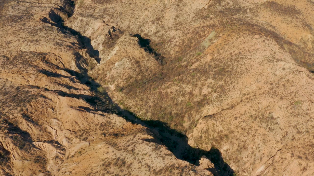 At 0:02, the diagonal riverbed divides pale tan ground from rougher brown rock textures in Cabo Pulmo, with scattered vegetation and no visible people.
