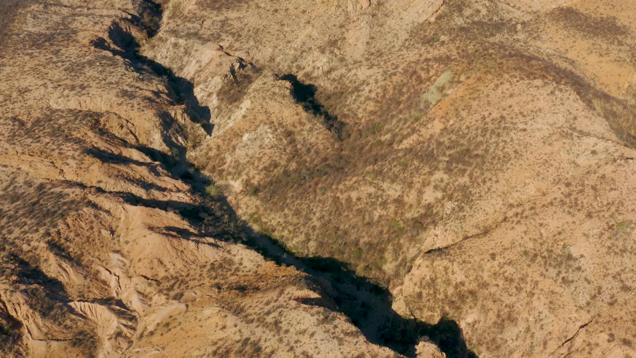 Aerial desert wash bordered by rugged tan ridges and dark ravine shadows.