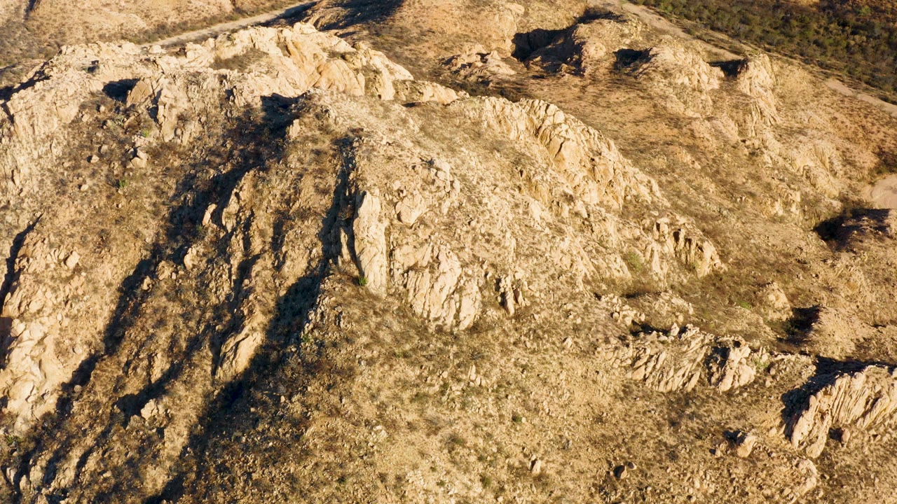 Aerial frame over a Cabo Pulmo rocky hillside with pale stone ridges, sparse dry grass, deep shadowed cracks, and no visible people.