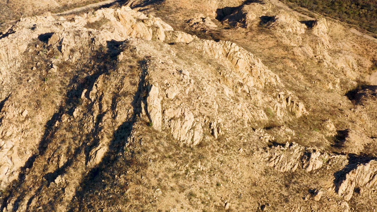 Aerial frame over a Cabo Pulmo hillside where pale stone bands cross dry brown grass and small shadow pockets, with no visible people.