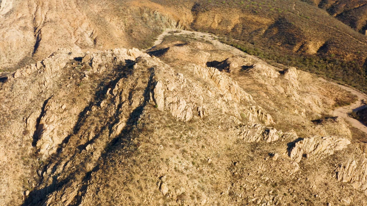 Aerial view in Cabo Pulmo, Baja California Sur with jagged tan rocks, dry grass, dark shadows, and a pale road curve near the upper right, no visible people.