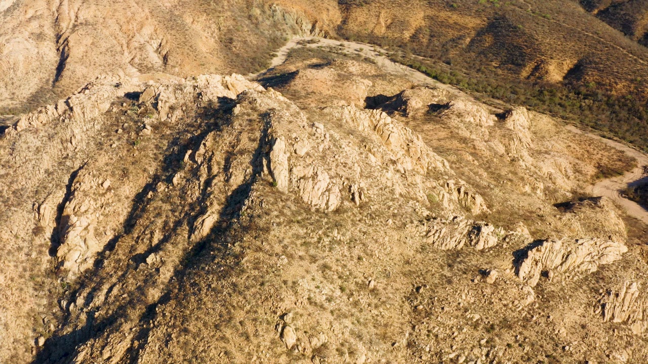 Sunlit aerial frame of Cabo Pulmo, Baja California Sur showing a broad rocky ridge, dry hillside textures, and a dirt road along the right background, no visible people.