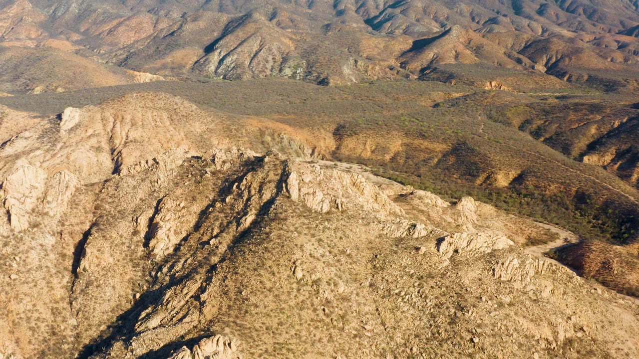 Aerial view in Cabo Pulmo with a pale rocky ridge dominating the foreground, muted green scrub on surrounding hills, a road curve at right, and no visible people.