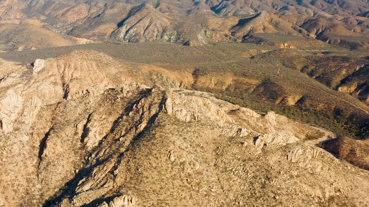 High-angle Cabo Pulmo frame showing sunlit tan ridgelines, darker gullies, low scrub, a road bend on the right side, and no visible people.