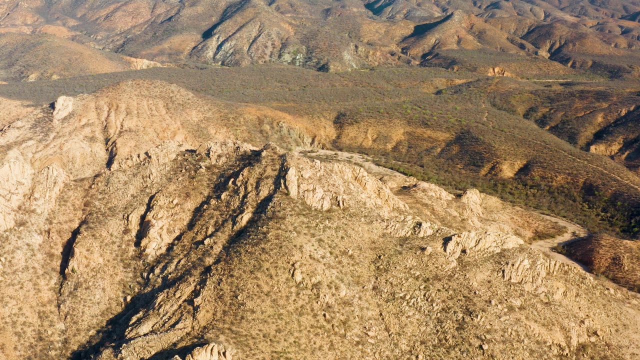 Cabo Pulmo aerial still with tan and ocher ridges across the foreground, scrubby midground hills, a winding road tracing the right side, and no visible people.