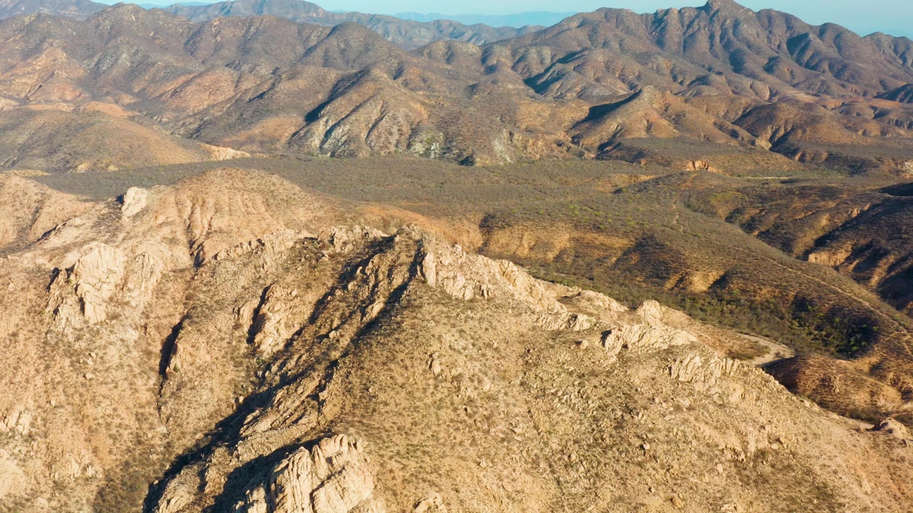 Wide elevated view in Cabo Pulmo showing a sunlit tan ridge in front, darker ravines through the midground, distant layered mountains, and no visible people.