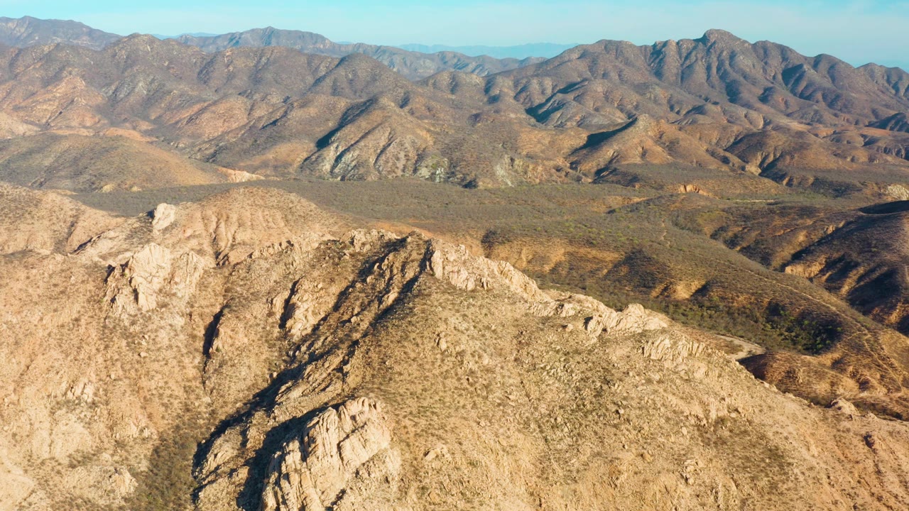 Aerial frame in Cabo Pulmo, Baja California Sur, showing the rocky ridge slightly farther across the frame, a muted green-brown valley floor, distant mountain ridges, and no visible people.