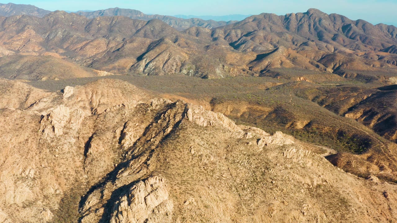 Mid-clip aerial frame over Cabo Pulmo mountain terrain, with tan fractured rock in the foreground, open dry valley in the middle distance, ridgelines across the horizon, and no visible people.