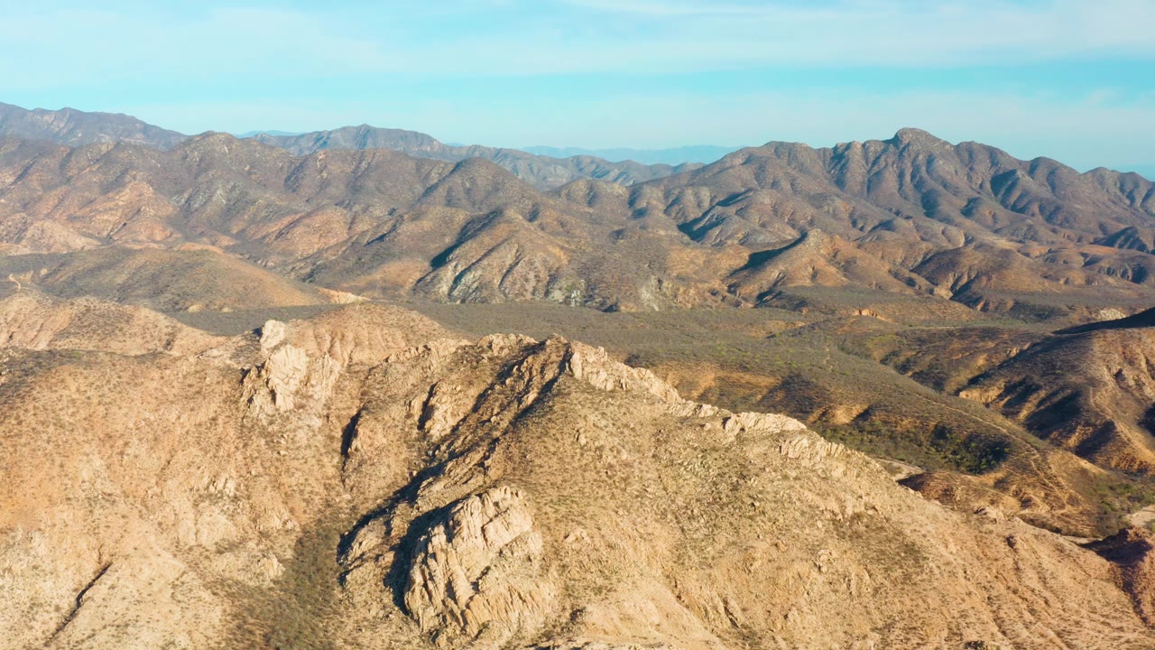 At 0:01, the Cabo Pulmo ridge view holds on the pale foreground rock, folded brown midground slopes, distant mountain line, clear blue sky, and no visible people.