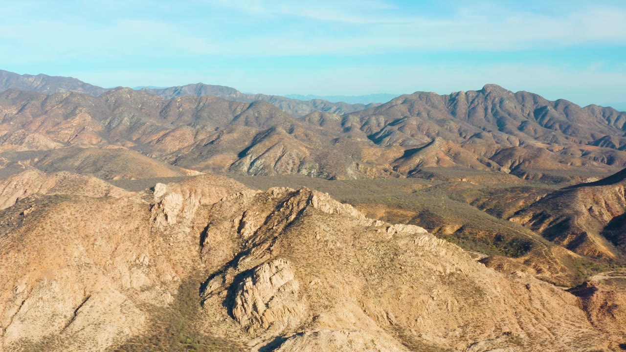 Elevated view over a tan rocky ridge toward layered Cabo Pulmo mountains, with dry valleys in the midground and no visible people.