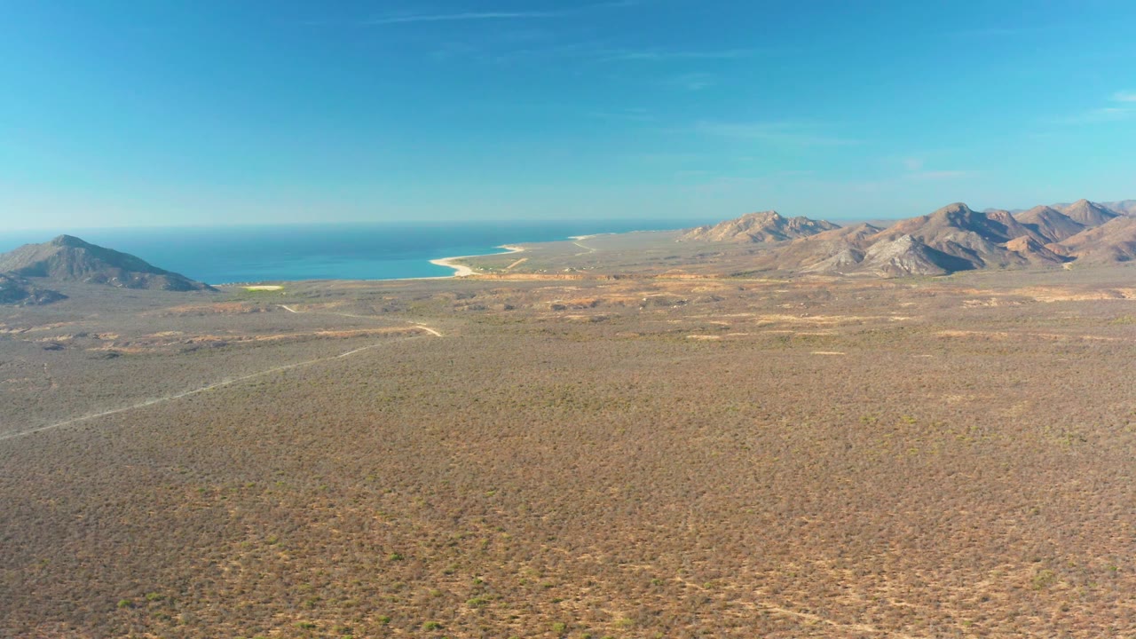 Aerial desert plain with mountains, ocean horizon, and pale shoreline.