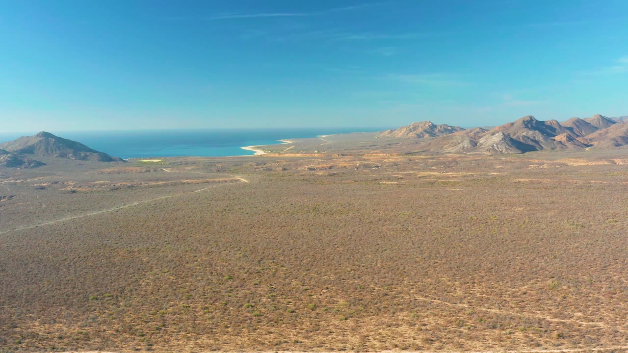 Aerial desert plain with distant mountains and ocean beyond a bright coastline.