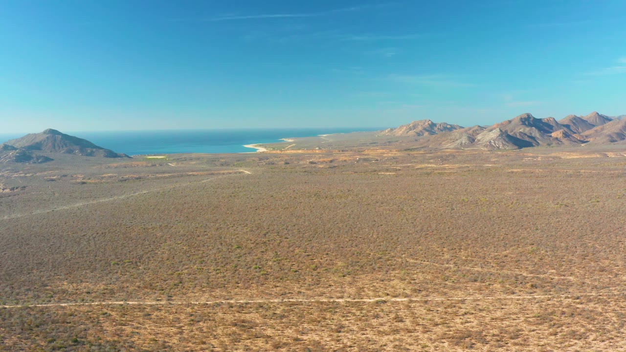 Cabo Pulmo aerial view with low desert vegetation filling the foreground, a thin road near the bottom edge, mountains in the distance, ocean beyond them, and no visible people.