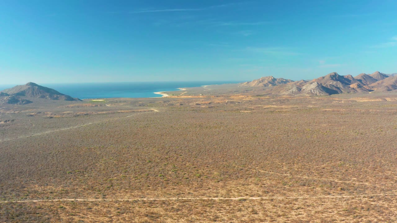 Mid-clip frame of Cabo Pulmo showing a high view over tan desert scrub, a pale track across the foreground, distant rocky mountains, blue ocean, and no visible people.
