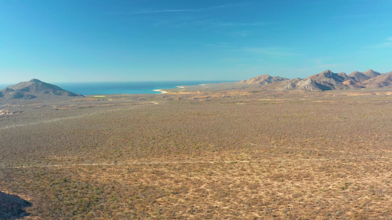 Opening aerial frame over Cabo Pulmo, Baja California Sur, showing scrubby desert plain leading to distant mountains, blue sea, and clear sky with no visible people.