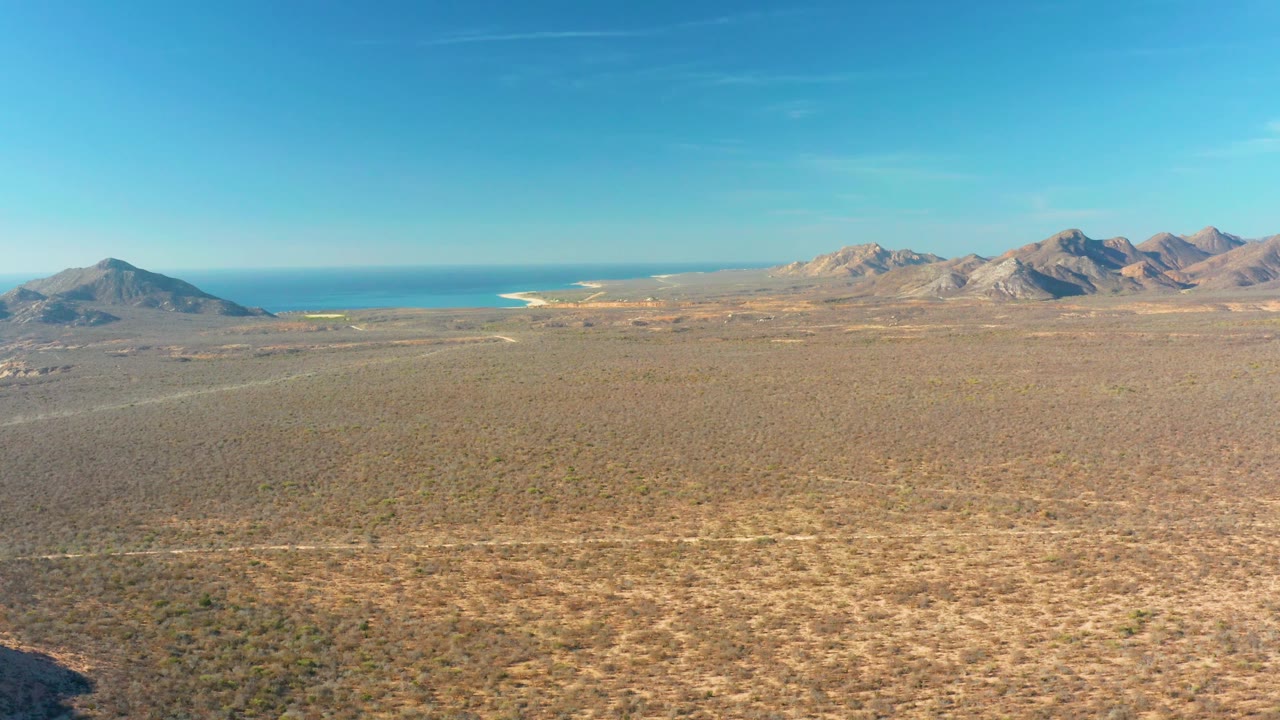 Cabo Pulmo aerial view with textured arid scrubland in the foreground, low mountain forms across the horizon, a strip of blue sea, and no visible people.