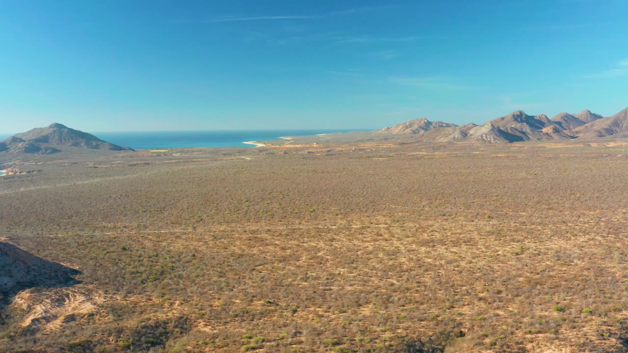 Opening frame over Cabo Pulmo desert plain with scrub in the foreground, mountains on both sides, blue ocean at the horizon, and no visible people.