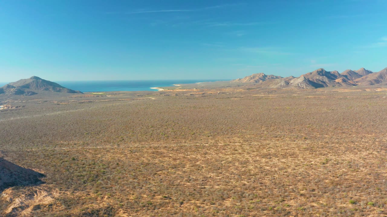 Mid-clip aerial frame over Cabo Pulmo scrubland, with a shadowed rocky slope entering from the lower left, ocean beyond the plain, and no visible people.
