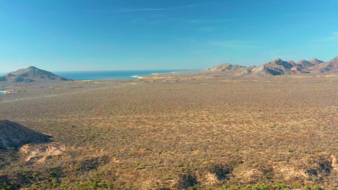 Wide Cabo Pulmo aerial frame with sparse desert vegetation across the plain, ocean on the horizon, mountains to the right, and no visible people.