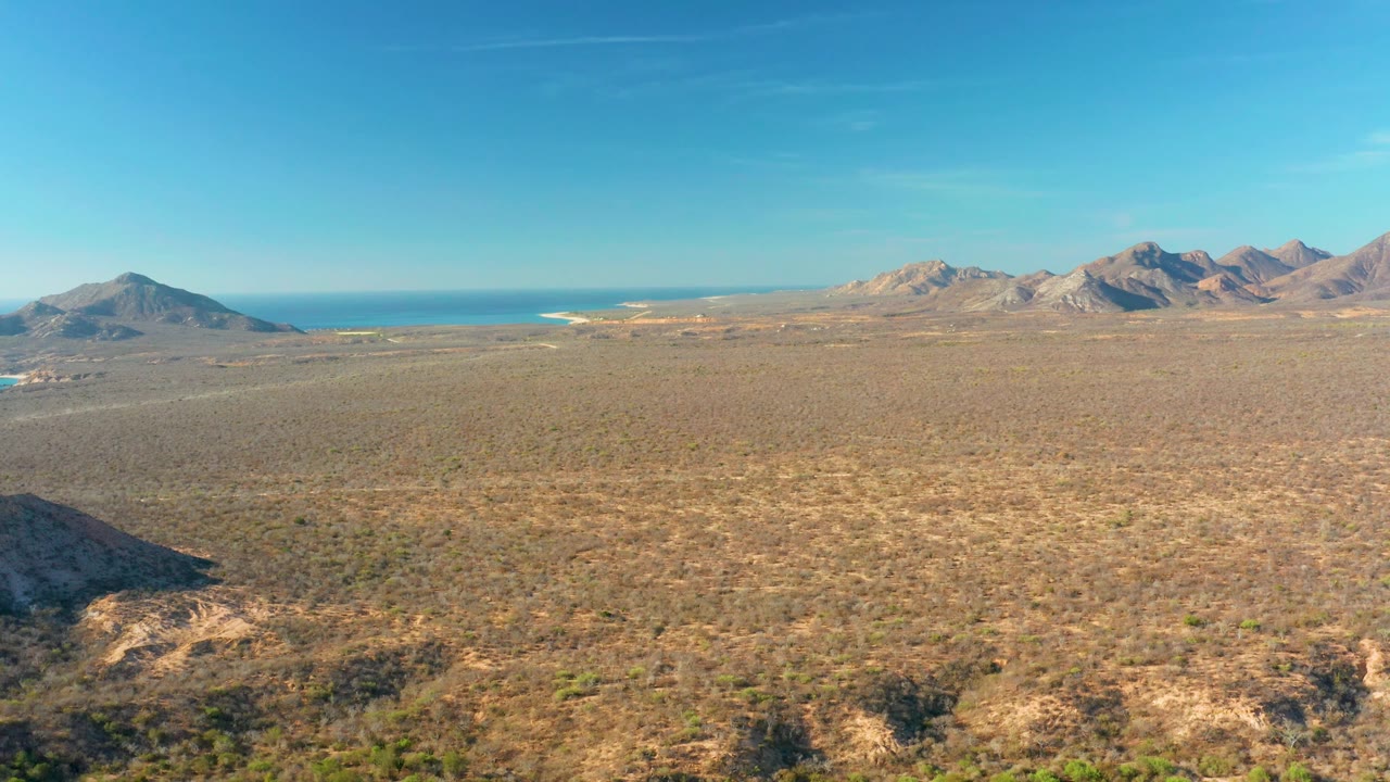 Cabo Pulmo desert plain seen from above with a small vehicle visible on the left side of the open terrain, backed by ocean, mountains, and no visible people.