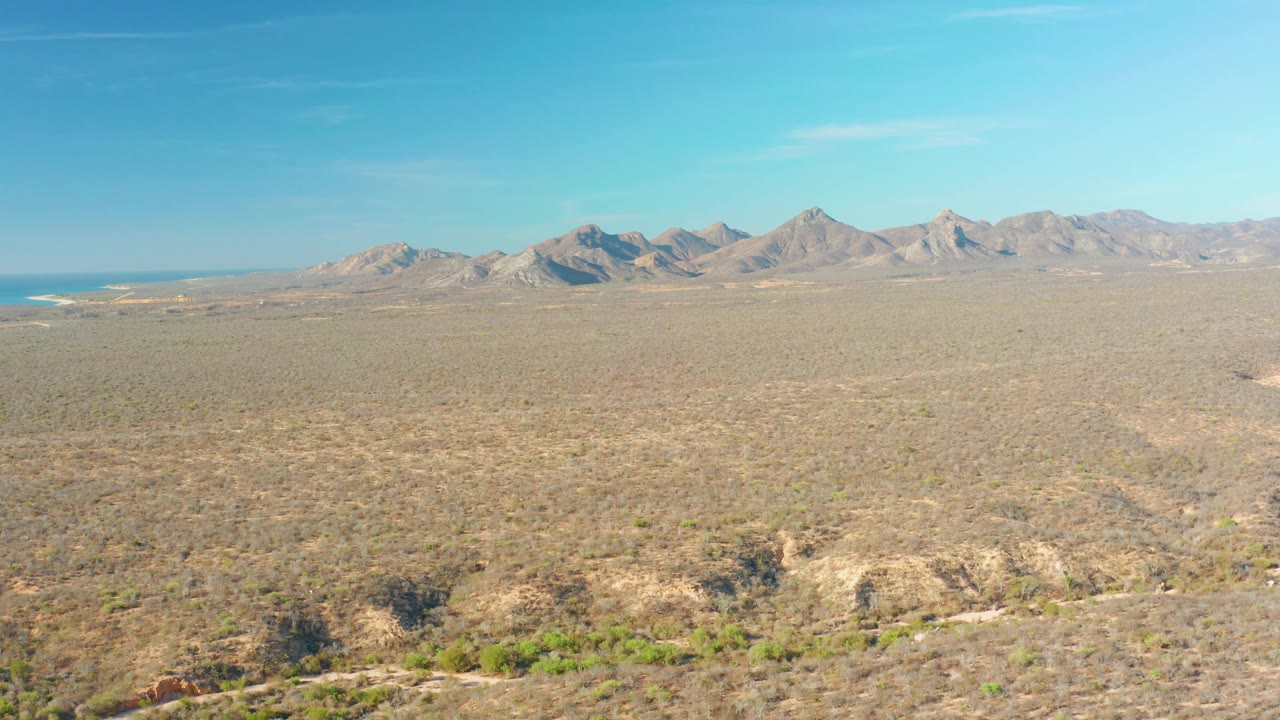 Open Cabo Pulmo desert plain extends to distant mountains, with a small blue coastal edge visible at the far left and no visible people.
