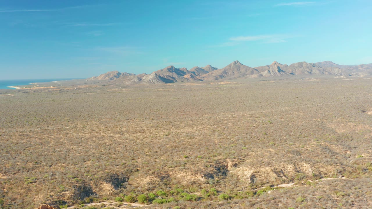 Elevated Cabo Pulmo view across tan scrub, shallow washes, and a continuous mountain range beneath a clear blue sky, with no visible people.