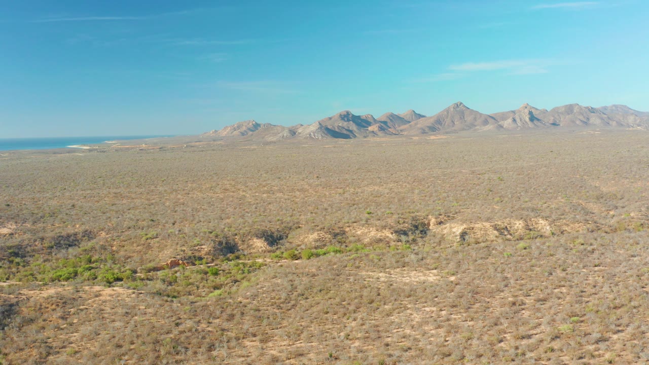 Opening frame of Cabo Pulmo desert plain with sparse brush, pale mountain range on the horizon, a small strip of blue water at far left, and no visible people.