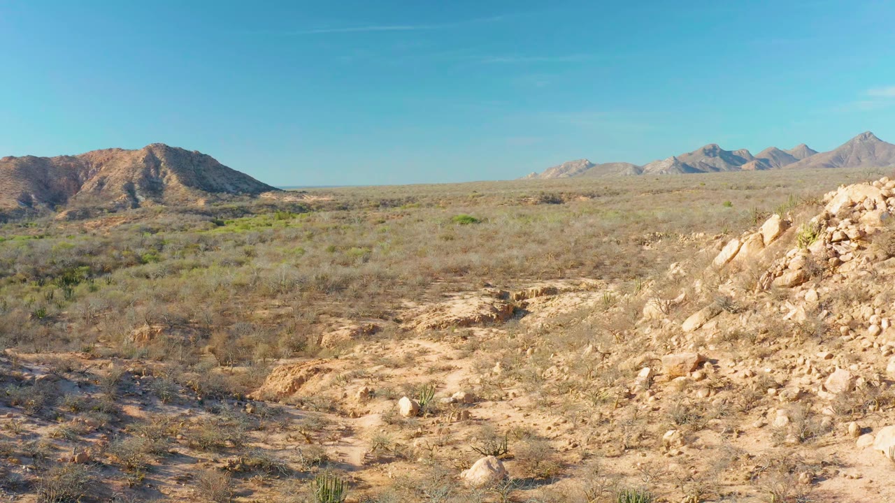 Cabo Pulmo desert valley at 0:01 with tan rocky ridges in the foreground, low scrub across the midground, mountain ridges in the background, blue sky, and no visible people.