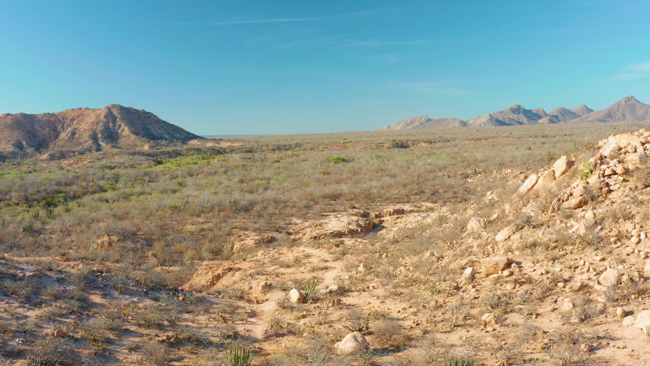 Cabo Pulmo, Baja California Sur, Mexico frame with a pale rocky slope filling the right foreground, open scrubland beyond it, distant mountain peaks, clear sky, and no visible people.