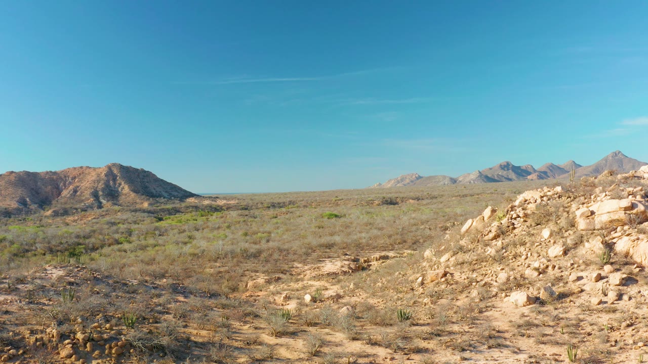 Opening frame of a Cabo Pulmo desert view with rocky foreground, sparse shrubs, distant mountain ridges, clear blue sky, and no visible people.