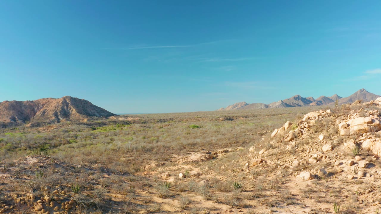 Rocky desert scrub with a boulder mound and distant hills under blue sky.
