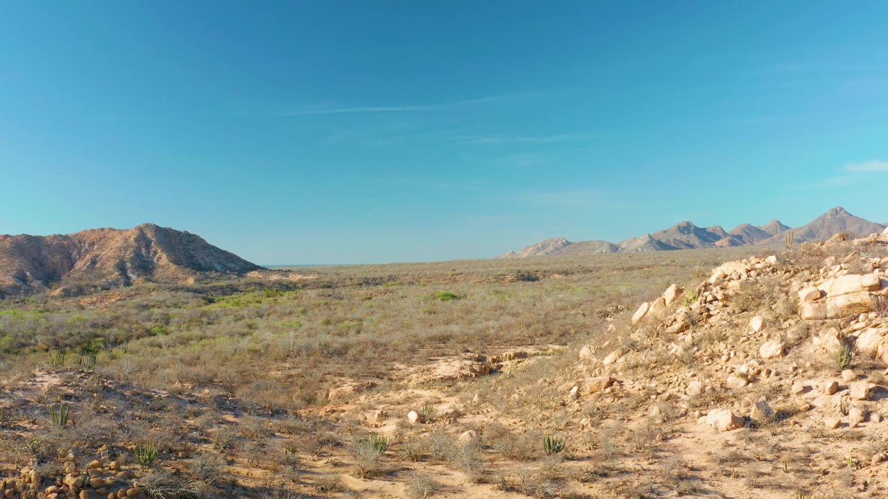 Cabo Pulmo landscape frame showing the rocky foreground mass shifted toward the right edge, open dry vegetation in the center, distant mountains, and no visible people.